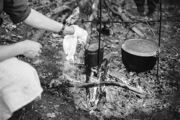 Re-enactor Dressed As World War II German Wehrmacht Soldier Cooking Food Over A Fire In An Old Marching Pot. Photo In Black And White Colors. Soldier Of WWII WW2 Times Re-enactor Dressed As World War II German Wehrmacht Soldier Cooking Food Over A Fire In An Old Marching Pot. Photo In Black And White Colors. Soldier Of WWII WW2 Times. fire letter b stock pictures, royalty-free photos & images