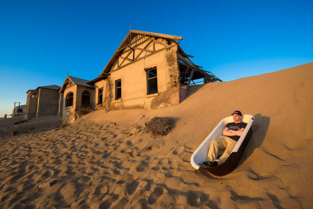 Tourist sits in a bathtub in Kolmanskop ghost town, Namibia Kolmanskop, Namibia - March 27, 2019 : Tourist sits in a bathtub placed outside in the sand desert around the ghost town of Kolmanskop. kolmanskop namibia stock pictures, royalty-free photos & images