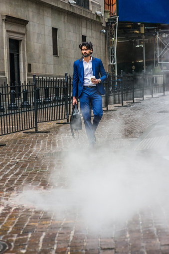 Raining day - grainy, foggy, wet feel. Young East Indian American Business Man with beard, wearing blue suit, white shirt, carrying leather hand bag, holding coffee cup, walking on street in New York.