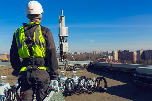 Professional industrial climber in helmet and uniform with telecomunication equipment in his hand and  antennas of GSM  DCS UMTS LTE bands, outdoor radio units  on the roof. Working process of upgrading telecommunication equipment