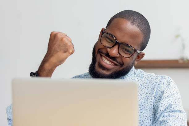 Smiling African American businessman celebrating success, using laptop Smiling African American businessman celebrating success, excited by great project results, using laptop, happy employee looking at screen, reading good news, business achievement concept sales win stock pictures, royalty-free photos & images