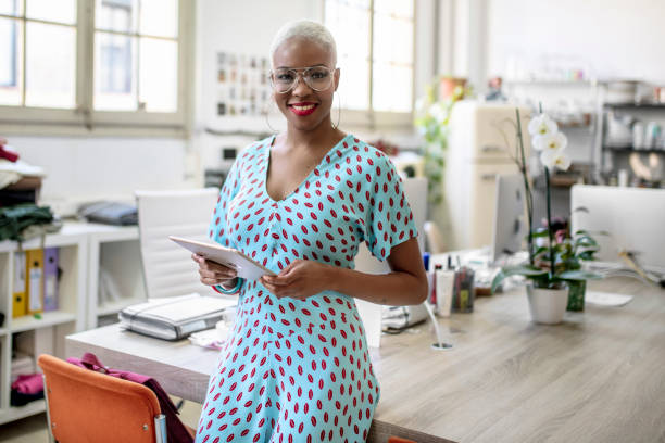 Portrait of smiling executive with digital tablet Portrait of smiling female designer with shaved head using digital tablet. Confident businesswoman is sitting on desk. She is wearing dress at creative office. shaved black women stock pictures, royalty-free photos & images