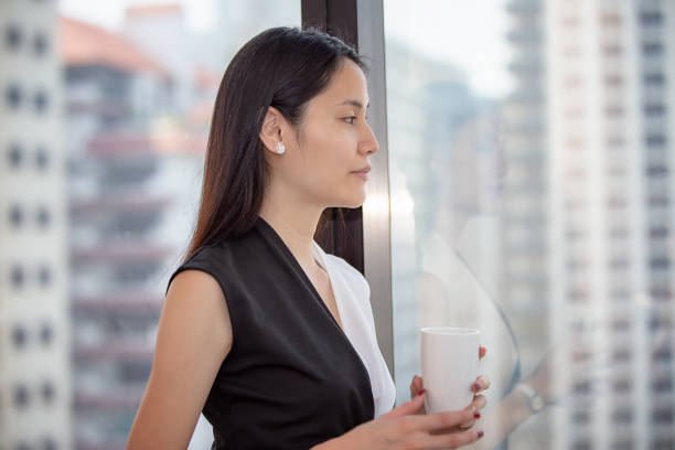 istock Businesswoman Looking Away While Holding Coffee Cup By Window In Office 1143877862