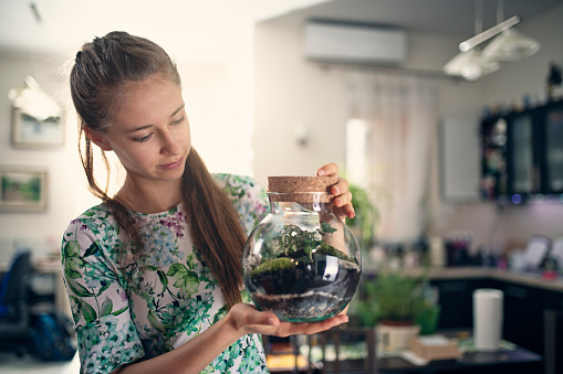 Teenage girl finished creating bottle garden at home. The girl is holding a jar of little plants inside - miniature living eco-system and beautiful home decoration.
Nikon D850
