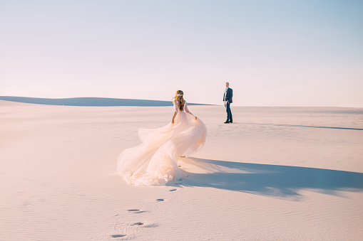 A woman runs to meet a man. Dress with a very long hairpin that flies in the wind. Photo from the back without a face. Background desert at sunset. Fine art photo.