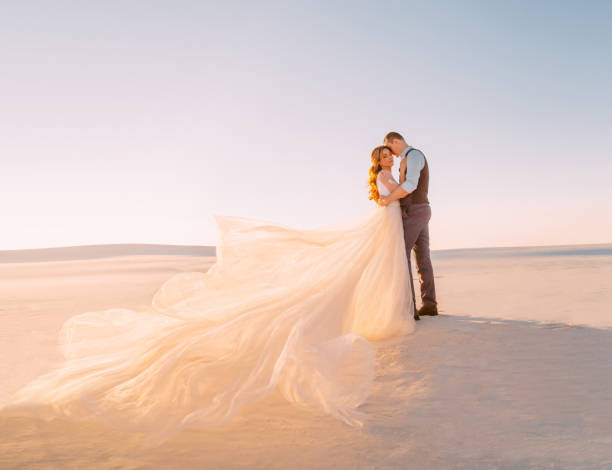Unusual wedding in the desert. A girl in a white dress ivory shade. Very long plume fluttering in the wind. A loving couple is embracing tenderly against the background of white sand and blue sky Unusual wedding in the desert. A girl in a white dress ivory shade. Very long plume fluttering in the wind. A loving couple is embracing tenderly against the background of white sand and blue sky. honeymoon photos stock pictures, royalty-free photos & images