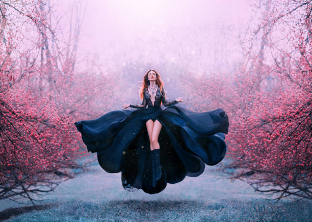 gorgeous red-haired hot woman in long flying black dress fluttering, witch turns into crow, dark queen in frozen spring flowering forest, levitation and flight, open breasts and bare sexy legs girl with bright red hair levitates above ground, powerful sorceress, forest goddess in black flying dress with lace on open chest and bare long legs, lady in flowering orange garden and hand raven. dark pink dress stock pictures, royalty-free photos & images