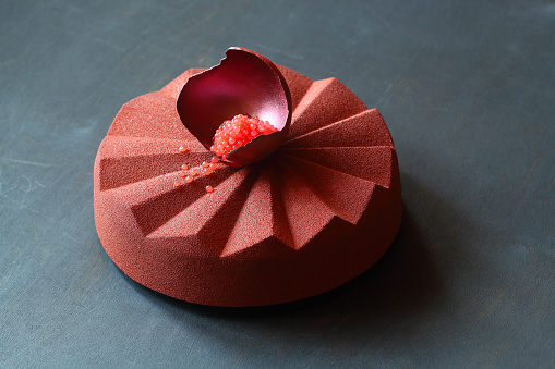 Contemporary Chocolate Mousse Cake made in geometric silicone mold, covered with brick color velvet spray and decorated with chocolate shell filled with blood orange caviar, on black background.