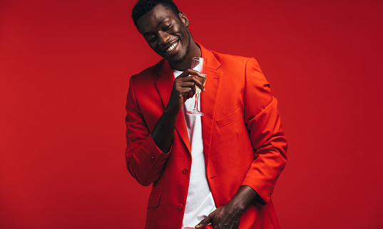 Cheerful young man in red jacket with a glass of champagne. Smiling african american man having a glass of champagne against red background.