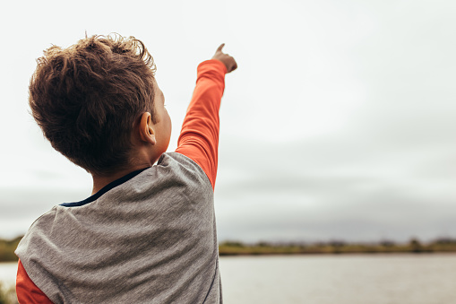 Rear view of a kid standing near the lake and showing something to someone. Close up back view of a boy pointing his finger to the sky.
