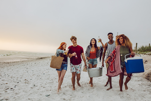 Group of young people walking on the beach carrying a cooler box and beverage tub. Young men and woman on sea shore.