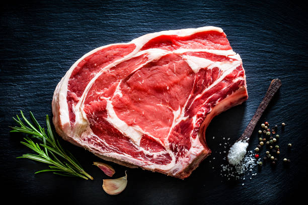 Raw rib steak shot from above on black background Top view of a thick juicy raw rib steak shot on black slate background. Ingredients for seasoning meat like rosemary, garlic, salt and pepper are included in the composition. Predominant colors are red and black. Low key DSRL studio photo taken with Canon EOS 5D Mk II and Canon EF 100mm f/2.8L Macro IS USM. beef chops stock pictures, royalty-free photos & images