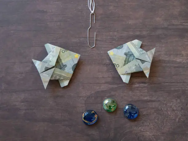 Origami fish made of five euro notes facing fishing-hook on dark wooden table area