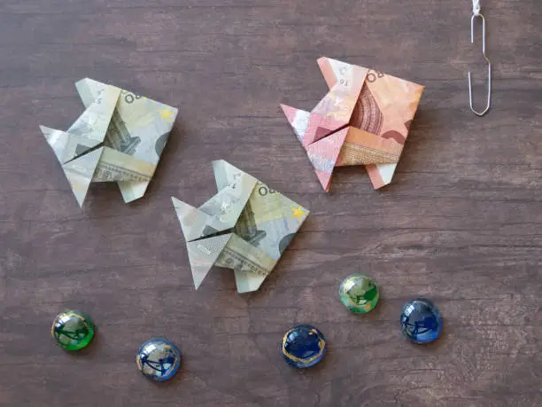 Three origami fish made of ten and five euro notes swimming towards fishing-hook on dark wooden table area