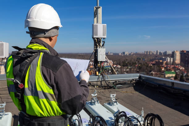 istock Professional industrial climber in helmet and uniform reads technical documentation and  antennas of GSM  DCS UMTS LTE bands, outdoor radio units  on the roof. Working process of upgrading telecommunication equipment. 1143976015