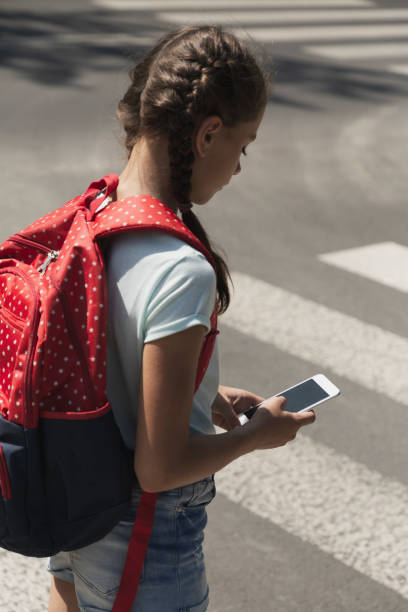 istock Girl with backpack using smartphone while walking through crosswalk to the school 1143981687
