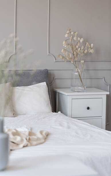 istock Bedside table with flowers in glass vase next to bed with white sheets 1143982941