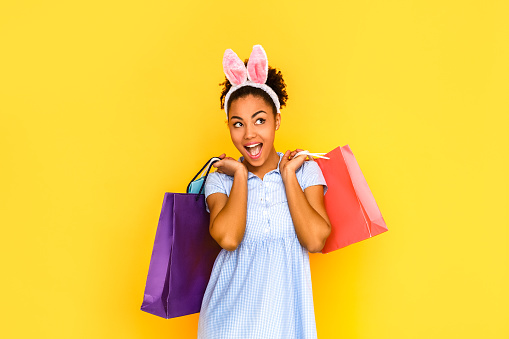 Young woman wearing cute dress and bunny ears headband standing isolated on yellow background holding shopping bags looking aside mouth opened smiling playful