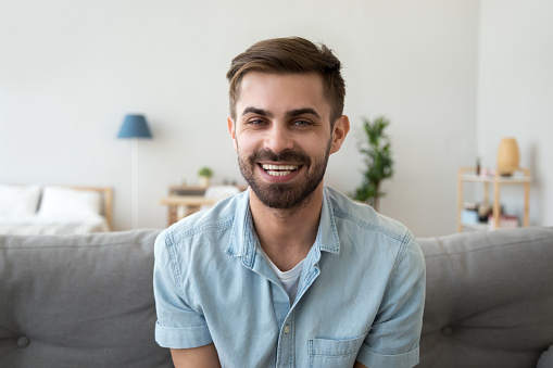 Head shot portrait of smiling happy man looking at camera, making video call, talking with friend, sitting on cozy coach at home, male vlogger recording webinar, communicate, broadcast live