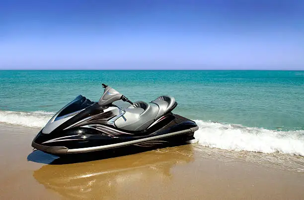 A black and silver jet ski waits invitingly on the beach with a beautiful aqua sea and clear blue sky as backdrop.