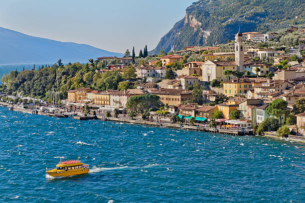 A view from a moving boat, where white foam cuts through the deep blue water of a lake, with mountains, hills, and a coastal town in the background. A tranquil summer travel scene capturing movement, freedom, and natural beauty, perfect for tourism, relaxation, adventure, and outdoor activities.