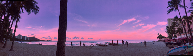 Colorful sunrise at Waikiki beach, Honolulu, Hawaii. There are people, surfboards and traditional polynesian boats on the edge. Panoramic view.