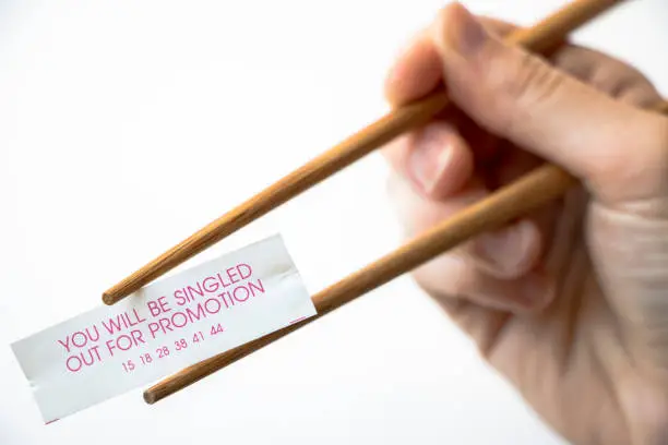 A paper fortune cookie fortune reading, 'You will be singled out for a promotion' being held by chopsticks and hand, white background.