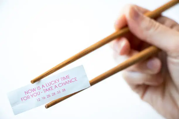 A paper fortune cookie fortune reading, 'Now is a lucky time for you - take a chance' being held by chopsticks and hand, white background.