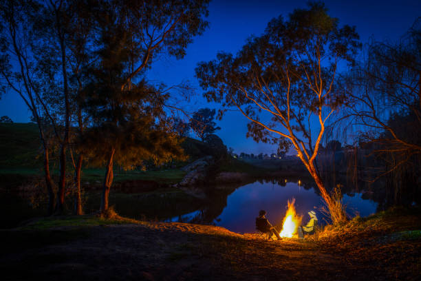 no crepúsculo duas pessoas sentam-se em uma fogueira na borda da água, new south wales, austrália - non urban scene standing water waterhole landscape - fotografias e filmes do acervo
