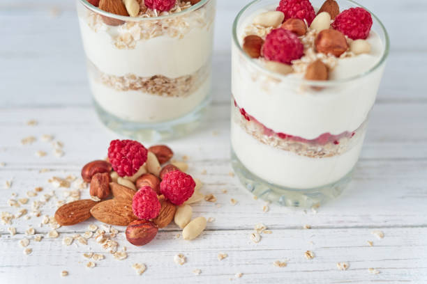 due bicchieri di muesli allo yogurt greco con lamponi, fiocchi di farina d'avena e noci su sfondo bianco. alimentazione sana - cashew apple fruit food jar foto e immagini stock