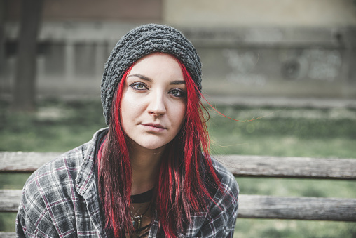 Young beautiful red hair girl sitting alone outdoors on the wooden bench with hat and shirt feeling anxious and depressed after she became a homeless person close up portrait
