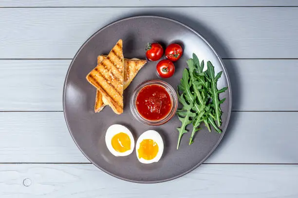 Photo of morning breakfast in a gray plate of boiled eggs, toast with cheese and vegetables on a white wooden background