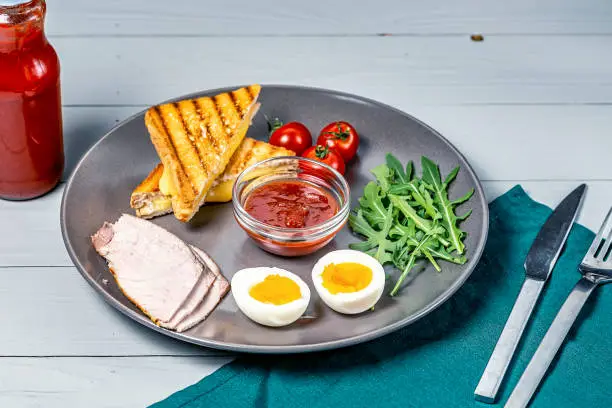 Photo of morning breakfast in a gray plate of boiled eggs, toast with cheese and vegetables on a white wooden background