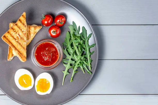 Photo of morning breakfast in a gray plate of boiled eggs, toast with cheese and vegetables on a white wooden background