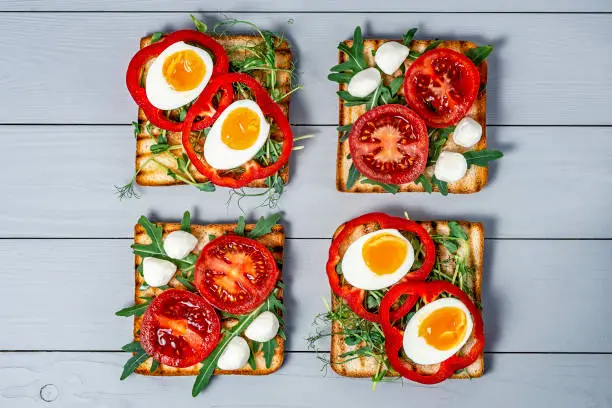 Photo of Sandwiches with fresh vegetables avocado with arugula and boiled eggs on fried toast with mozzarella cheese on a gray wooden background with a place for copy space, flat lay