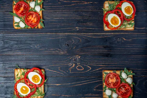 Photo of sandwiches with fresh vegetables avocado arugula and boiled eggs on fried toast with mozzarella cheese on a black wooden background with a place for copy space, flat lay