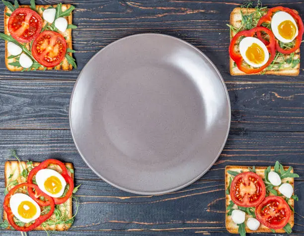 Photo of sandwiches with fresh vegetables avocado with arugula and boiled eggs on fried toast with mozzarella cheese on a black wooden background with an empty plate