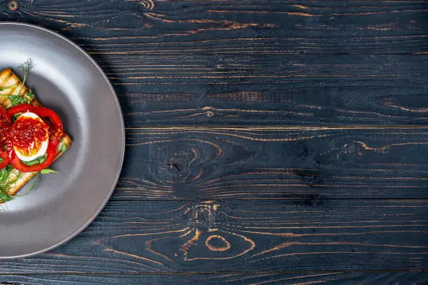 Photo of Sandwich with fresh vegetables and boiled eggs on roasted toast on a gray plate on a black wooden background with a place for copy space