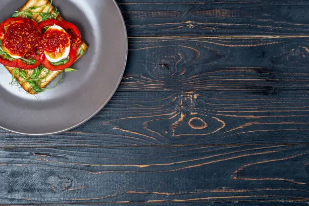 Photo of Sandwich with fresh vegetables and boiled eggs on roasted toast on a gray plate on a black wooden background with a place for copy space