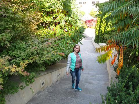Long-haired young adult girl in autumn clothes walks up the stairs, top view. A woman in jeans, a jacket and sneakers is walking upstairs in a park between green and yellow trees