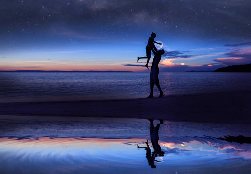 Mirror reflection of Silhouette father and daughter on the beach with million stars galaxy ,the moon and blue sky early morning.