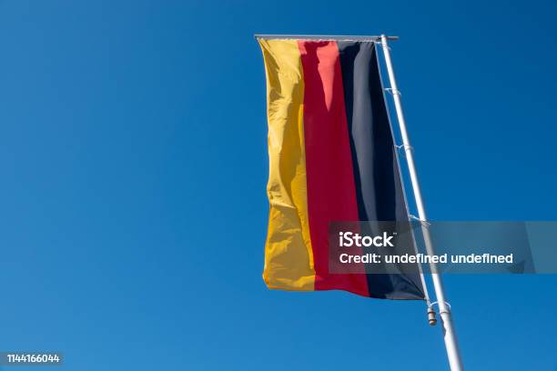 A German Flag Blows In Front Of A Blue Sky Stock Photo - Download Image Now - Allegory Painting, Architecture, Backgrounds