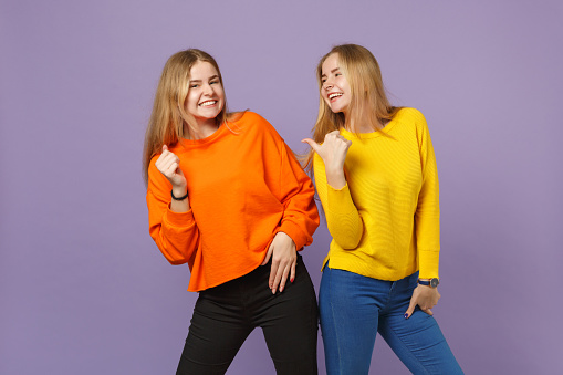 Two laughing joyful young blonde twins sisters girls in vivid colorful clothes pointing thumb aside isolated on pastel violet blue wall background. People family lifestyle concept. Mock up copy space