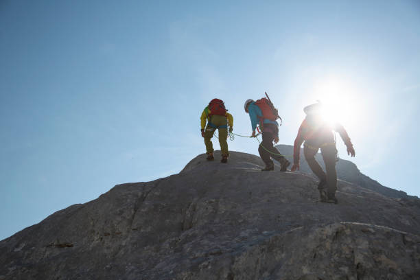 Group of mountaineers walking on the edge Three mountain climbers silhouettes against a blue sky with sun. group mountain silhouette stock pictures, royalty-free photos & images