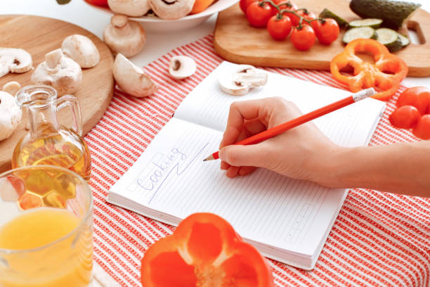 Hand writes a recipe. Many vegetables on the table and a notebook Hand writes a recipe. Many vegetables on the table and a notebook with an inscription cooking. Indoor, studio shoot, kitchen interior. Indoor, studio shoot, kitchen interior finely stock pictures, royalty-free photos & images