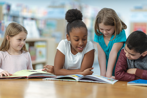 A group of friends are sitting at a table in their classroom. They are reading a book together as they smile and laugh.