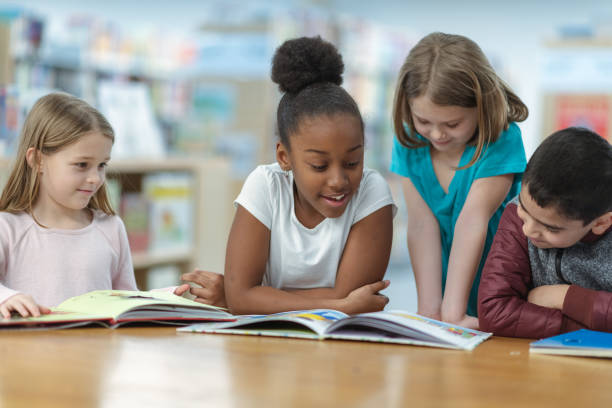 Children Reading A group of friends are sitting at a table in their classroom. They are reading a book together as they smile and laugh. 8 9 years stock pictures, royalty-free photos & images