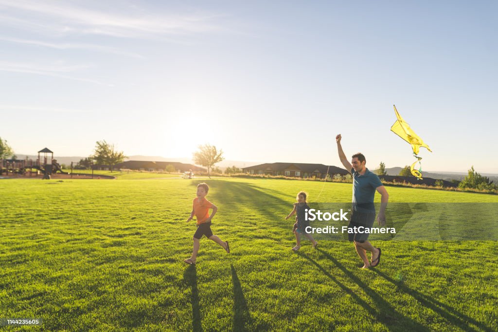 Dad flying kites with his kids at the park Dad and his two kids fly a kite through a grassy field on a beautiful summer day. He's helping hold it aloft while the youngest, a girl, flies it. Big brother is running in front. There is a playground in the background. Public Park Stock Photo
