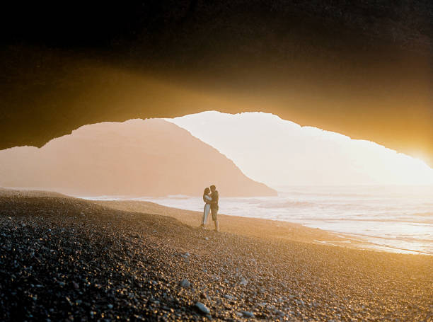 Young couple enjoying the sunset. Romantic couple standing near natural huge arch on the beach. Handsome man and attractive woman enjoying sunset in Morocco. moroccan girl stock pictures, royalty-free photos & images