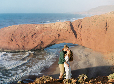 Young couple in love having fun on sunset view for huge natural arch in the ocean in Morocco.  Wanderlust, freedom and lifestyle concept.